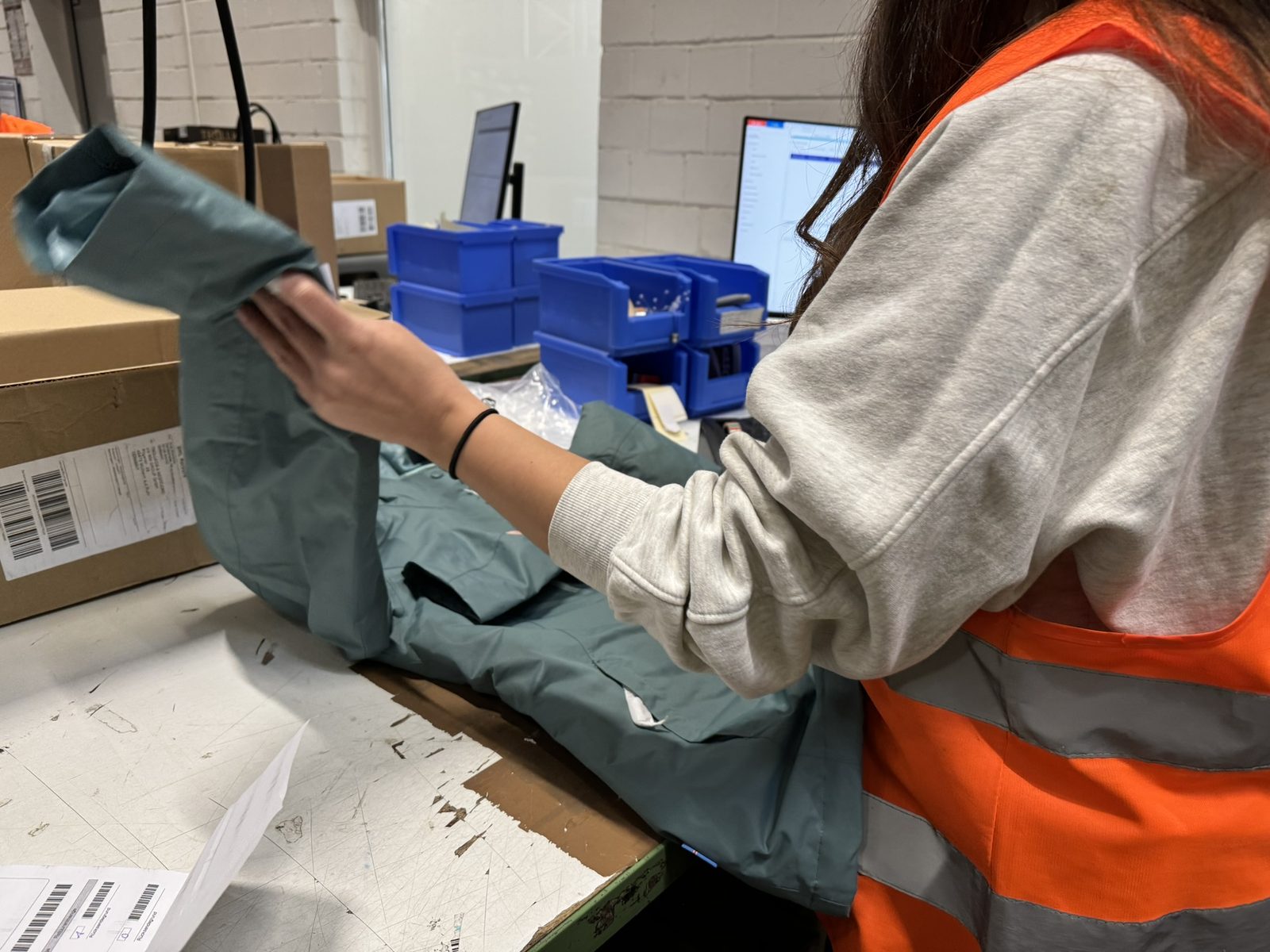An employee folding a garment at the packing station, next to a shipping carton and a screen.