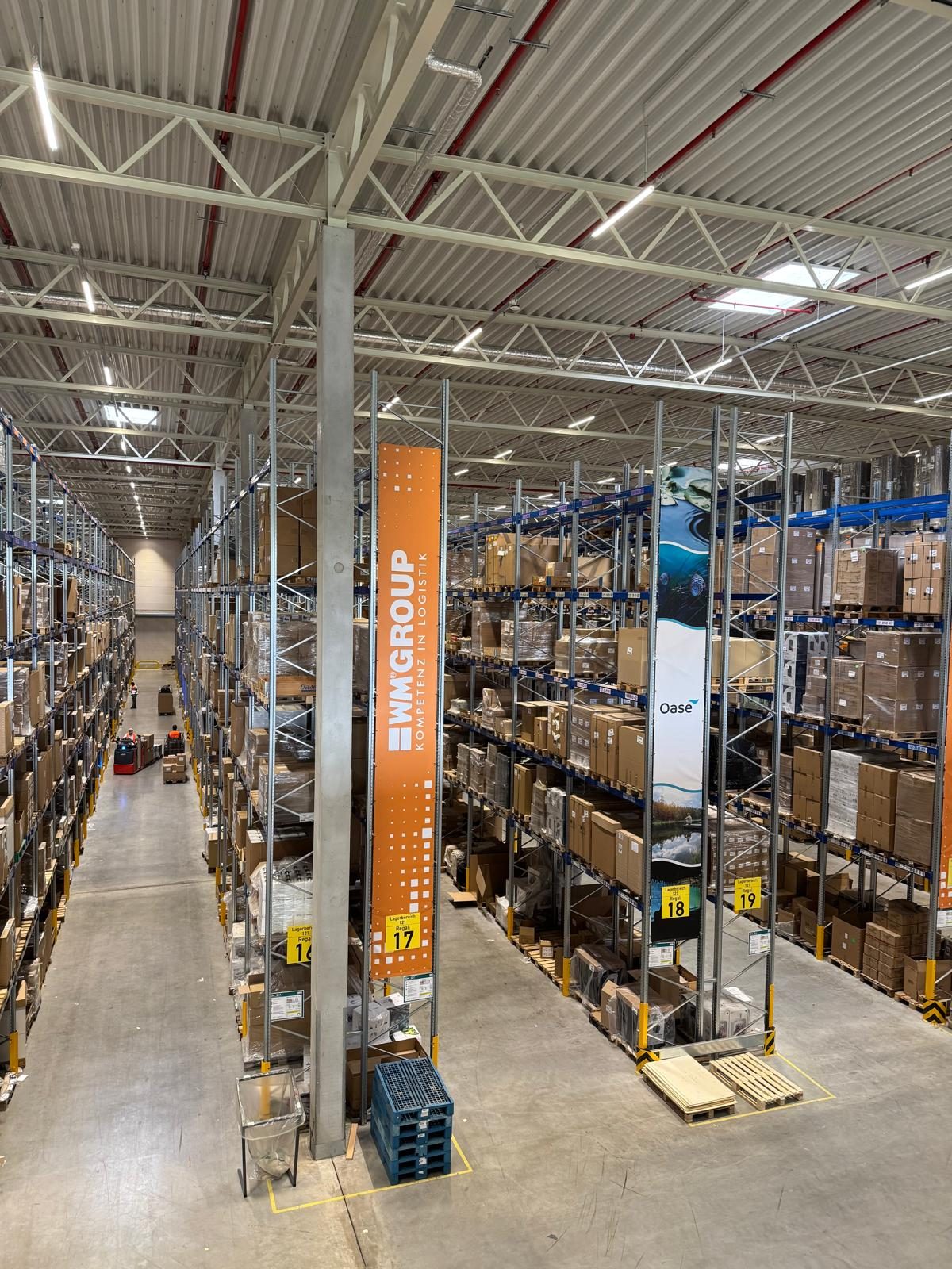 A warehouse aisle between two racking rows, an orange WM GROUP banner at the end.
